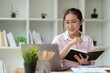© Phimwilai - Office worker or businesswoman, attractive Asian woman in eyeglasses researching Analyze information about the business plan. take notes in a book Notebook while using a laptop computer at her desk.