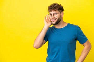 Wall Mural - Young handsome caucasian man isolated on yellow background listening to something by putting hand on the ear