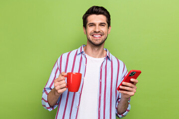 Fototapete - Photo of satisfied person with bristle wear stylish clothes hold cup of tea chatting on smartphone isolated on green color background