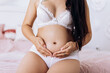 Pregnant woman strokes hugging the belly tummy abdomen enjoying pregnancy sitting on bed dressed underwear. Future family, baby infant expecting child inside
