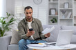 © Tetiana - African american man working at home with documents, worried sitting on sofa at home and reading letter he received, looking at bills, calling to solve problems