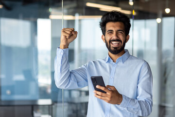 Indian young male businessman standing in office and using phone, looking at screen and happy with success. Shows victory gesture and celebration with hands to the camera. Got good news.
