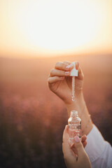 Close up female hands holding essential oil in a bottle with pipette in lavender field at sunset.