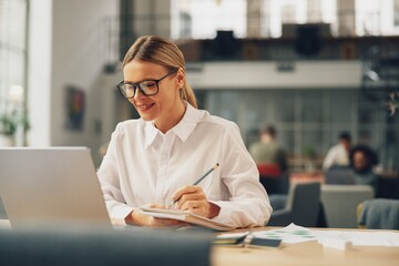 Wall Mural - Woman office worker working on laptop and making notes while sitting in coworking