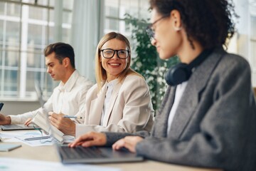 Wall Mural - Smiling woman manager working on laptop in modern coworking while sitting near colleagues