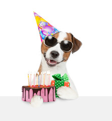 Poster - Jack russell terrier puppy wearing sunglasses and party cap holds gift box and birthday cake with lot of candle above empty white board. isolated on white background