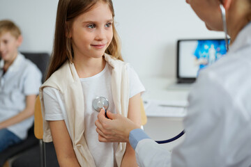 Wall Mural - Doctor and kid patient are in the clinic. Physician in white coat examining a smiling young girl with a stethoscope, close up. Medicine, therapy concept