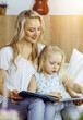 © rogerphoto - Happy family. Blonde young mother reading a book to her cute daughter while sitting at wooden floor in sunny room. Motherhood concept