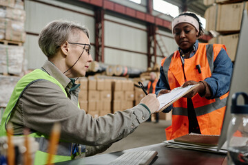 Foreman sitting at workplace with computer and signing document on receipt of the goods giving by worker