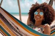 © igolaizola - Young african american woman with afro hairstyle and sunglasses relaxing in hammock on tropical beach.