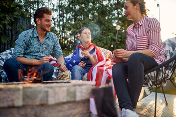 Joyful and happy family of man, woman and girl sitting outdoors by a fireplace roasting marshmallows enjoying sunny summer day together.