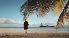 Woman Walking Barefoot White Sand Beach At Sunset. Tourist Girl In Black Dress Enjoy Tropical Island Landscape, Blue Ocean Water And Coconut Palms. Travel, Tourism, Holiday. Back View, Slow Motion
