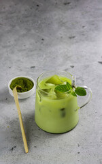 A cup of antioxidant matcha drink made of ground powder of green tea leaves with milk, ice cubes and fresh mint on serving tray with matcha spoon and matcha powder bowl. Iced matcha latte. Copy space