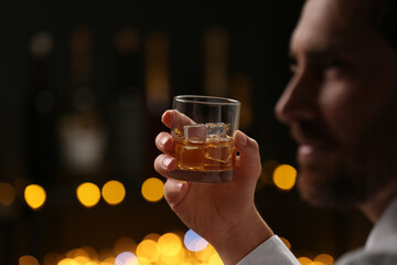 Canvas Print - Man holding glass of whiskey with ice cubes against blurred lights, selective focus