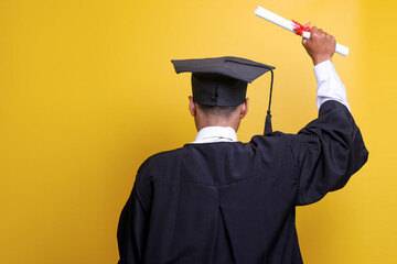 Education and graduation concept. Rear view of a young male graduate with a gown and a cap raising his arms as a celebration after getting a university certificate during a college graduation