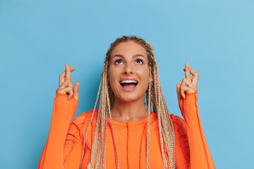 Wall Mural - Close-up shot of pretty woman wearing orange longsleeve top on blue background, smiling and crossing her fingers for luck, happy time concept, copy space