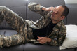 © Angelov - Unhappy man in camouflage uniform sitting at arm chair over white studio background, touching his head, soldier suffering from posttraumatic stress disorder, returning back home after military service