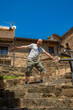 © sirbouman - Bald man with half a beard smiling with open mouth, expressive eyes and arms outstretched to give a cheerful and funny surprise standing on a stone staircase in a rural village on a sunny day