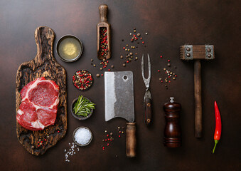 Wall Mural - Meat cleavers,tenderizer,fork,knife and other kitchen meat utensils with oil,herbs,salt and pepper on dark background with raw rib eye steak fillet.