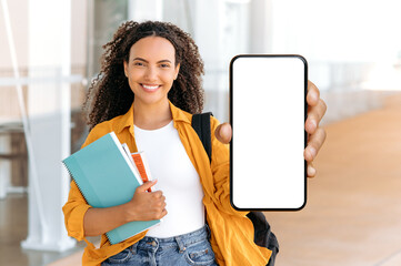 Copy-space, mock-up. Positive curly brazilian or hispanic female student, with a backpack, books and notebooks in hand, stand near university, shows smart phone with white blank mock-up screen, smile
