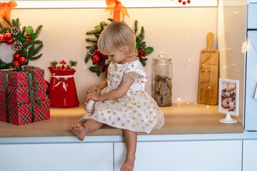 Christmas kitchen girl. Portrait of a charming little girl 2 years old, she sits on a table in a white dress and drinks from a red mug in a decorated kitchen for christmas.