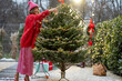 Young woman in red decorates lush Christmas tree with festive ballls and garland at backyard of her house on snow fall, preparing for a winter holidays