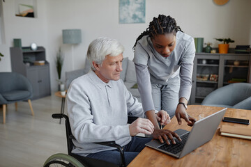 Portrait of senior man with disability learning to use computer with nurse or caretaker assisting