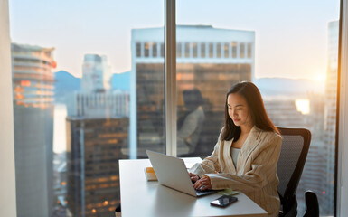 Young busy Asian business woman executive working on laptop in corporate office. Professional businesswoman marketing sales manager using computer technology sitting at table, city view from window.