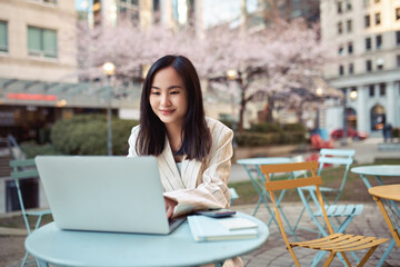 Wall Mural - Young happy Asian business woman professional or student sitting at street table using laptop hybrid working or remote learning online typing on computer technology in city park.