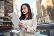 © insta_photos - Young busy pretty Asian business woman professional office manager executive or global international company employee standing on big city street outside using digital tablet fintech pad device.