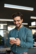 Happy Young Latin Business Man Executive, Businessman Manager Standing In Office Holding Smartphone Using Mobile Cell Phone Managing Digital Apps On Cellphone At Work. Vertical