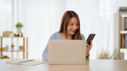 Young asian woman happy working on computer laptop and hand typing chat message on mobile smartphone at home. Video call, Online meeting