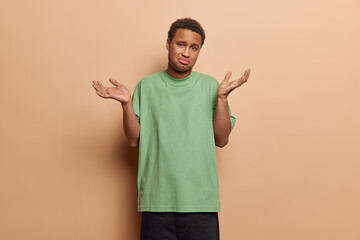 Wall Mural - People emotions concept. Studio photo of young confused African male not knowing how to solve difficult problem or task wearing casual clothes standing isolated in centre on beige background