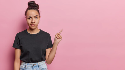 Serious displeased Latin woman with hair bun raises eyebrows looks attentively at camera purses lips has dimple on cheek dressed in casual black t shirt and jeans isolated over pink background