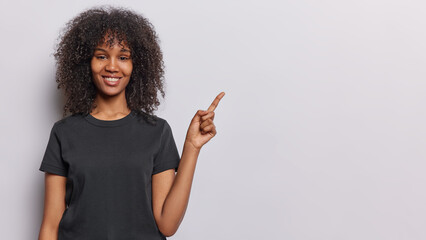 People positive emotions concept. Studio waist up of young cheerful smiling broadly African girl standing on left isolated on white background wearing casual black tshirt pointing at promotion space