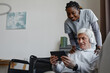 © Seventyfour - Portrait of white haired senior man with disability relaxing at home and using tablet with nurse assisting, copy space