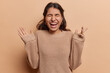 © wayhome.studio  - Waist up shot of emotional dark haired Iranian girl keeps hands raised up exclaims loudly from joy keeps eyes closed dressed in knitted sweater isolated over brown background. Human emotions