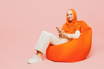 Wall Mural - Full body happy fun young arabian asian muslim woman wears orange abaya hijab sit in bag chair hold use mobile cell phone isolated on plain pink background. Uae middle eastern islam religious concept.