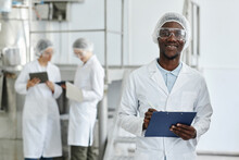 Waist Up Portrait Of Black Young Man Wearing Lab Coat And Smiling At Camera In Clean Workshop Of Pharmaceutical Factory, Copy Space
