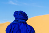 Fototapeta  - Close-up. Background. Berber in blue traditional clothing against the backdrop of a large dune. View from behind. Tunisia, Africa