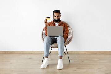 Wall Mural - Indian man holding credit card and laptop shopping against wall