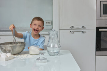 Funny child in the kitchen with white furniture learns to knead the dough in a metal bowl. The happy boy is confidently controlled by kitchen appliances while making a cake