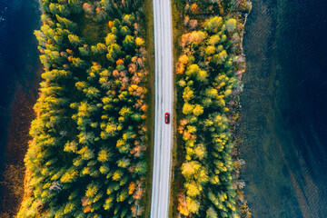 Aerial view of road through fall woods with blue water lakes.  Red car with a roof rack on a country road in Finland.