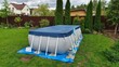 A gray rectangular frame swimming pool stands on the grassy lawn of the infield and is covered with an awning. Nearby thuja and flowers grow in flower beds The pool is made of PVC material