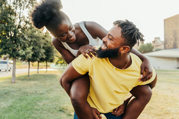 Young happy smiling black african couple in love having fun in the park. Boyfriend holds piggyback his girlfriend at sunset. Summer vacations, lifestyle concept.