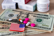 A Medical Cost, Big Pharma Concept With A Glass Of Water, Various Medicine, US Dollar Bills And The American Flag.