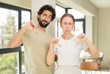 Wall Mural - young adult couple looking sad, disappointed or angry, showing thumbs down in disagreement, feeling frustrated