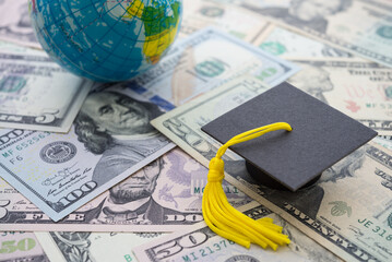 Graduation cap or hat on US dollars banknotes and globe world background. Abroad education, global international study business concept. Congratulation to get university degree or master.