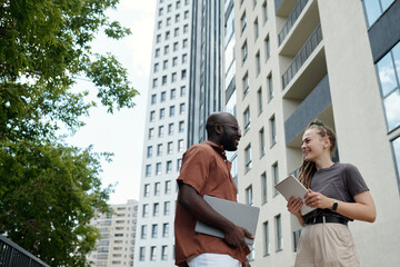 Wall Mural - Happy young intercultural white collar workers in casualwear holding wireless gadgets while having discussion of working points outdoor