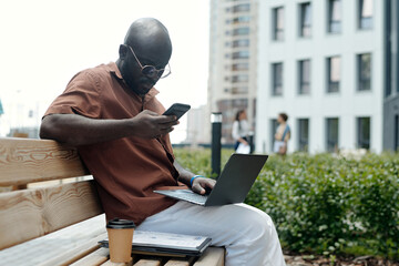 Fototapete - Young busy serious broker texting in smartphone and using laptop while sitting on bench in urban environment against modern building